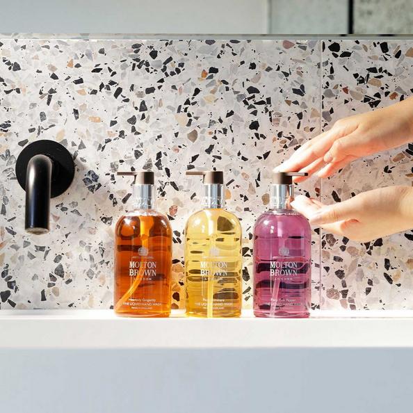 An image of three bottles of Molton Brown Hand Wash on a sink and a persons hands